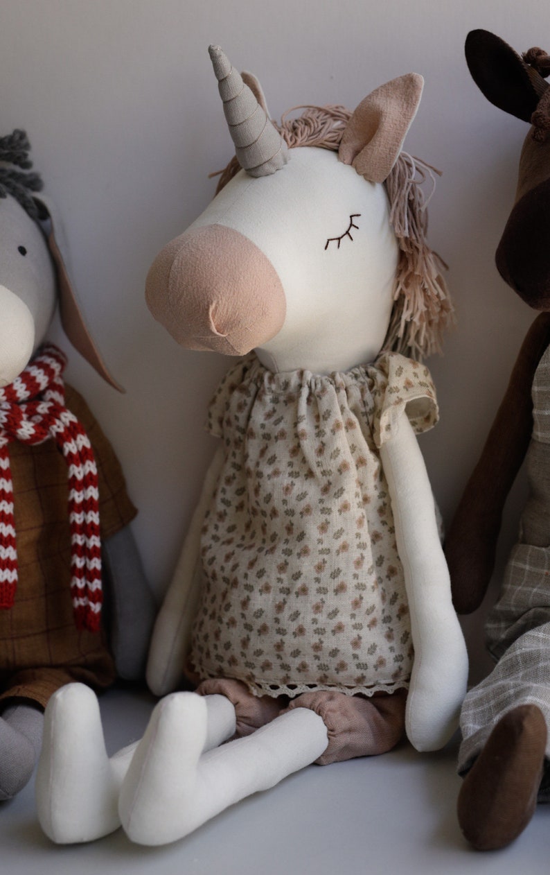 Puede incluir: Un juguete de peluche de unicornio blanco con la nariz rosa y los ojos cerrados. El unicornio lleva un vestido floral marr&oacute;n y blanco y pantalones marrones. El unicornio est&aacute; sentado en una superficie gris.