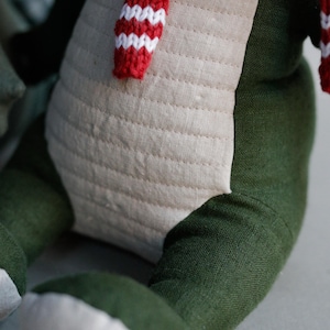 May include: A plush toy with a green body and white legs, wearing a red and white striped scarf. The toy is made of soft fabric and has a quilted design on the front.