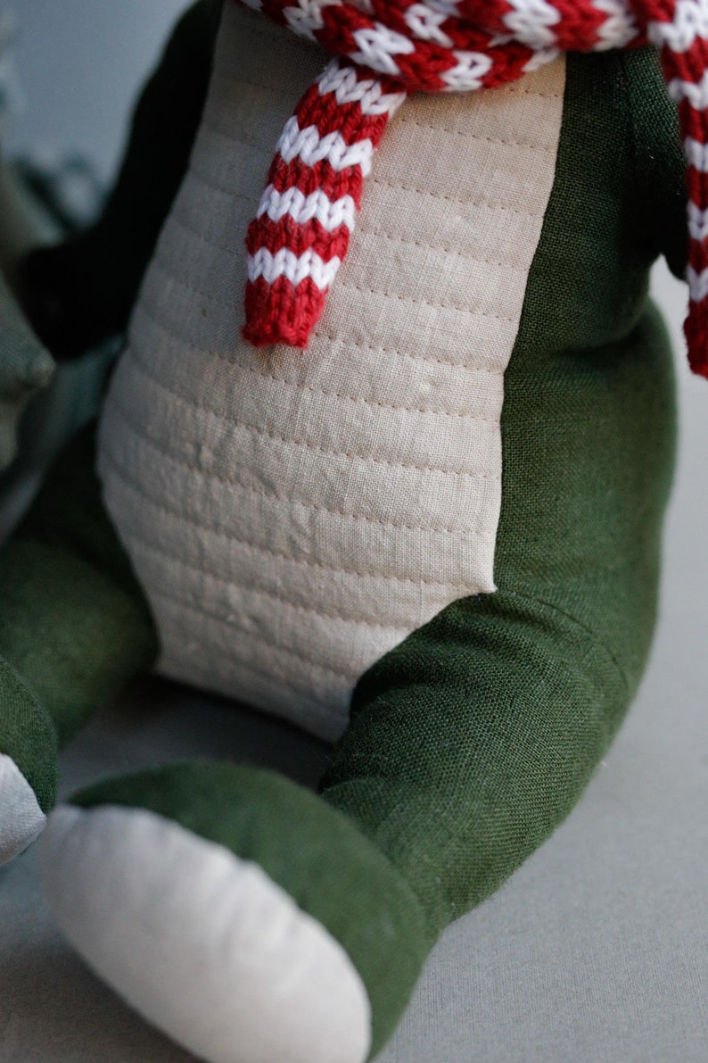 May include: A plush toy with a green body and white legs, wearing a red and white striped scarf. The toy is made of soft fabric and has a quilted design on the front.