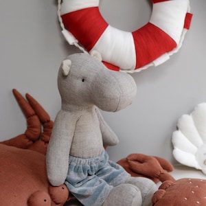 May include: A gray stuffed hippopotamus wearing blue denim shorts sits on a brown stuffed crab with a red and white life preserver in the background.