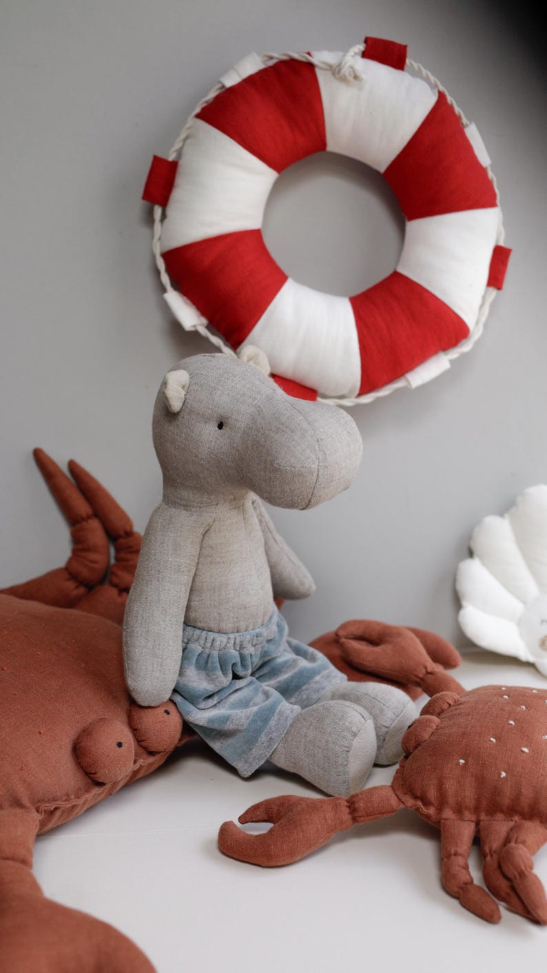 May include: A gray stuffed hippopotamus wearing blue denim shorts sits on a brown stuffed crab with a red and white life preserver in the background.