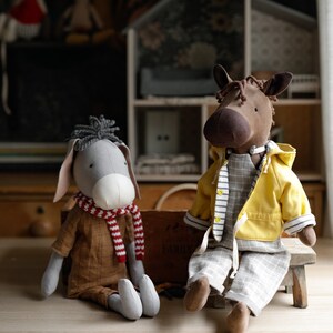 Puede incluir: Dos animales de peluche, un burro gris con una bufanda roja y blanca y un caballo marr&oacute;n con una chaqueta amarilla y pantalones grises, sentados en un taburete de madera.