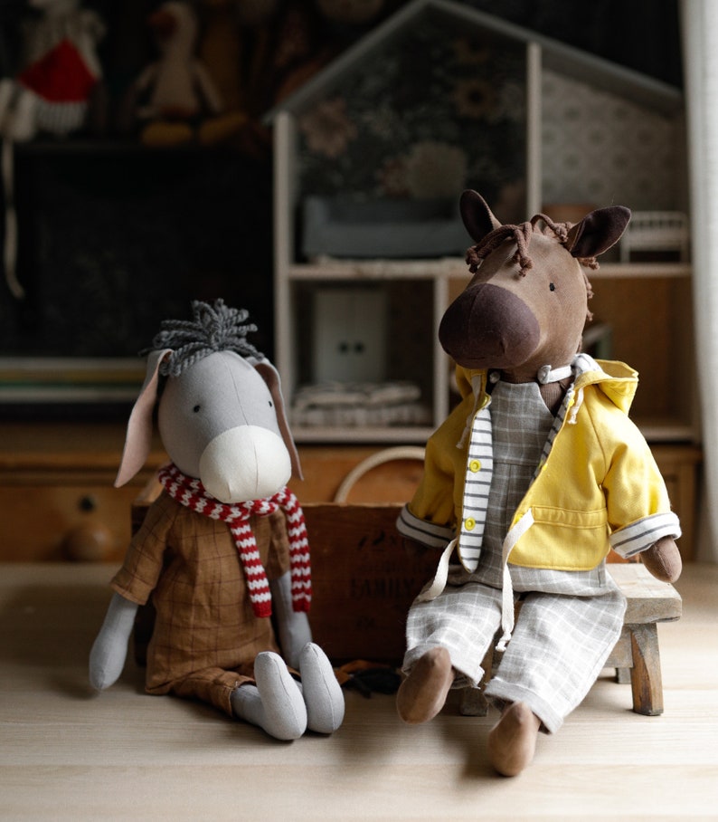 Puede incluir: Dos animales de peluche, un burro gris con una bufanda roja y blanca y un caballo marr&oacute;n con una chaqueta amarilla y pantalones grises, sentados en un taburete de madera.