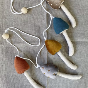 Puede incluir: Una guirlanda de cinco setas de fieltro en diferentes colores y patrones, incluyendo marr&oacute;n, gris, azul, amarillo y gris con puntos dorados. Las setas est&aacute;n unidas a una cuerda blanca con cuentas de madera en cada extremo.