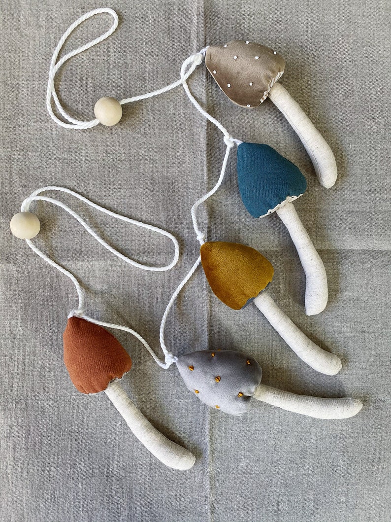 Puede incluir: Una guirlanda de cinco setas de fieltro en diferentes colores y patrones, incluyendo marr&oacute;n, gris, azul, amarillo y gris con puntos dorados. Las setas est&aacute;n unidas a una cuerda blanca con cuentas de madera en cada extremo.