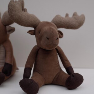 May include: A brown plush moose toy with large, floppy ears and a brown nose. The moose is sitting with its legs crossed and has a soft, fuzzy texture.