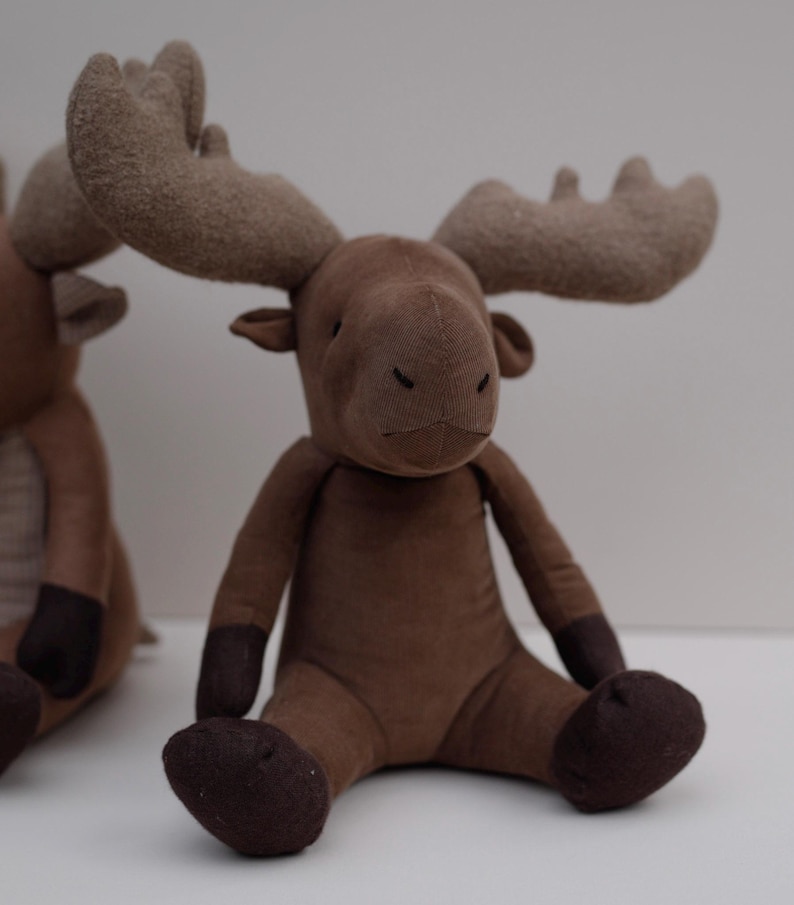 May include: A brown plush moose toy with large, floppy ears and a brown nose. The moose is sitting with its legs crossed and has a soft, fuzzy texture.