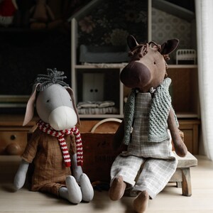 Puede incluir: Dos animales de peluche, un burro gris y un caballo marr&oacute;n, est&aacute;n sentados sobre una superficie de madera. El burro lleva una bufanda a rayas rojas y blancas y una camisa marr&oacute;n. El caballo lleva una bufanda de punto gris y una camisa a cuadros.