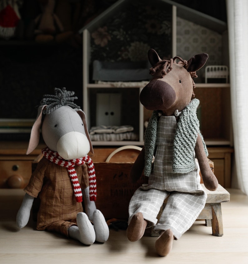 Puede incluir: Dos animales de peluche, un burro gris y un caballo marr&oacute;n, est&aacute;n sentados sobre una superficie de madera. El burro lleva una bufanda a rayas rojas y blancas y una camisa marr&oacute;n. El caballo lleva una bufanda de punto gris y una camisa a cuadros.