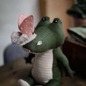 May include: A green plush crocodile toy with a pink and white butterfly on its head. The crocodile is sitting on a wooden surface.
