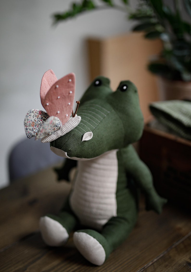 May include: A green plush crocodile toy with a pink and white butterfly on its head. The crocodile is sitting on a wooden surface.