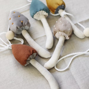 Puede incluir: Una guirnalda de cinco setas de fieltro en varios colores, incluyendo gris, marr&oacute;n, azul verdoso, dorado y beige. Las setas est&aacute;n enhebradas con un cord&oacute;n blanco y tienen tallos blancos.