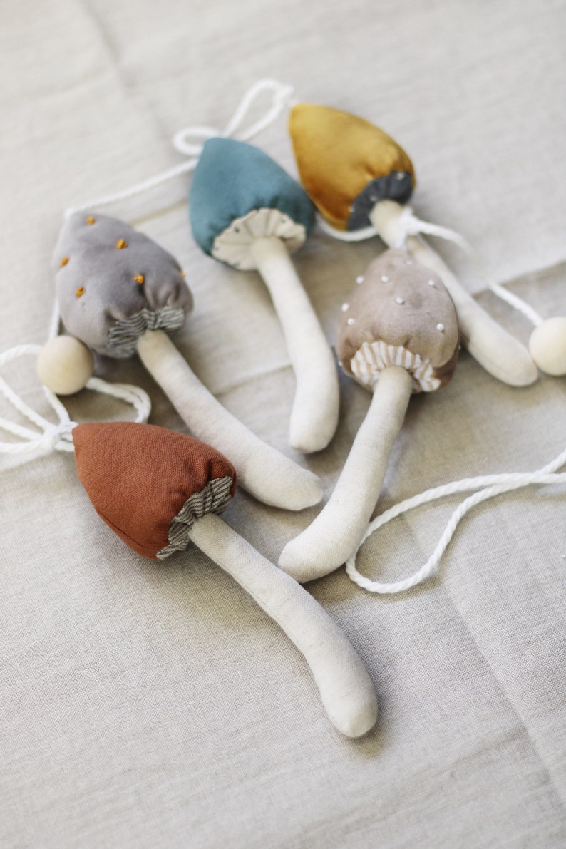Puede incluir: Una guirnalda de cinco setas de fieltro en varios colores, incluyendo gris, marr&oacute;n, azul verdoso, dorado y beige. Las setas est&aacute;n enhebradas con un cord&oacute;n blanco y tienen tallos blancos.
