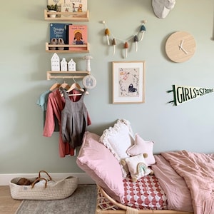 Puede incluir: Una pared verde claro con un estante de madera que contiene libros y una figurita de casa blanca de cer&aacute;mica. Un reloj de madera blanco con manecillas negras est&aacute; en la pared. Una cabeza de animal de madera blanca est&aacute; en la pared. Una cama de madera con una manta rosa y una almohada blanca est&aacute; en la habitaci&oacute;n. La cama tiene una s&aacute;bana con estampado rojo y blanco. El texto "GIRLS are STRONG" est&aacute; en la pared.
