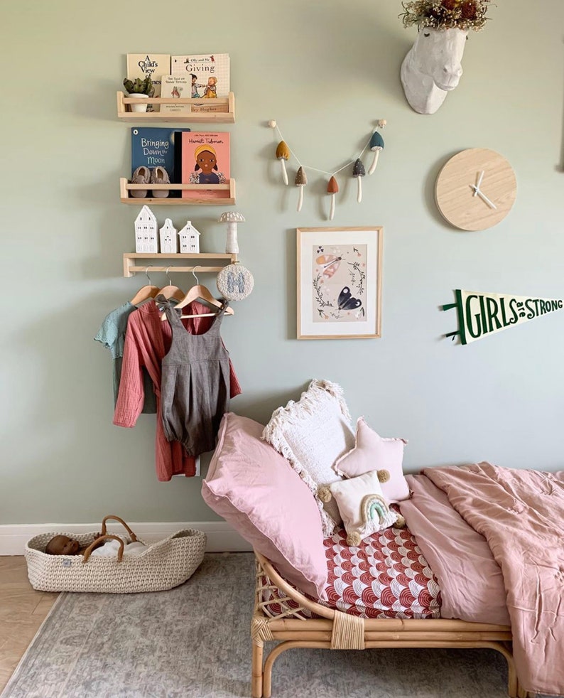 Puede incluir: Una pared verde claro con un estante de madera que contiene libros y una figurita de casa blanca de cer&aacute;mica. Un reloj de madera blanco con manecillas negras est&aacute; en la pared. Una cabeza de animal de madera blanca est&aacute; en la pared. Una cama de madera con una manta rosa y una almohada blanca est&aacute; en la habitaci&oacute;n. La cama tiene una s&aacute;bana con estampado rojo y blanco. El texto "GIRLS are STRONG" est&aacute; en la pared.