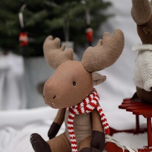 May include: A brown plush moose with large antlers wearing a red and white striped scarf. The moose is sitting on a white surface with a red sled in the background.