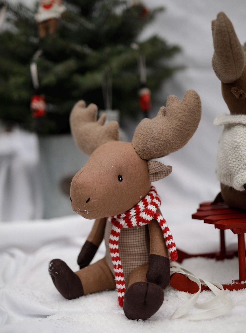 May include: A brown plush moose with large antlers wearing a red and white striped scarf. The moose is sitting on a white surface with a red sled in the background.