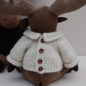May include: A brown plush moose wearing a white knitted cardigan with brown buttons. The moose has large antlers and a brown face.