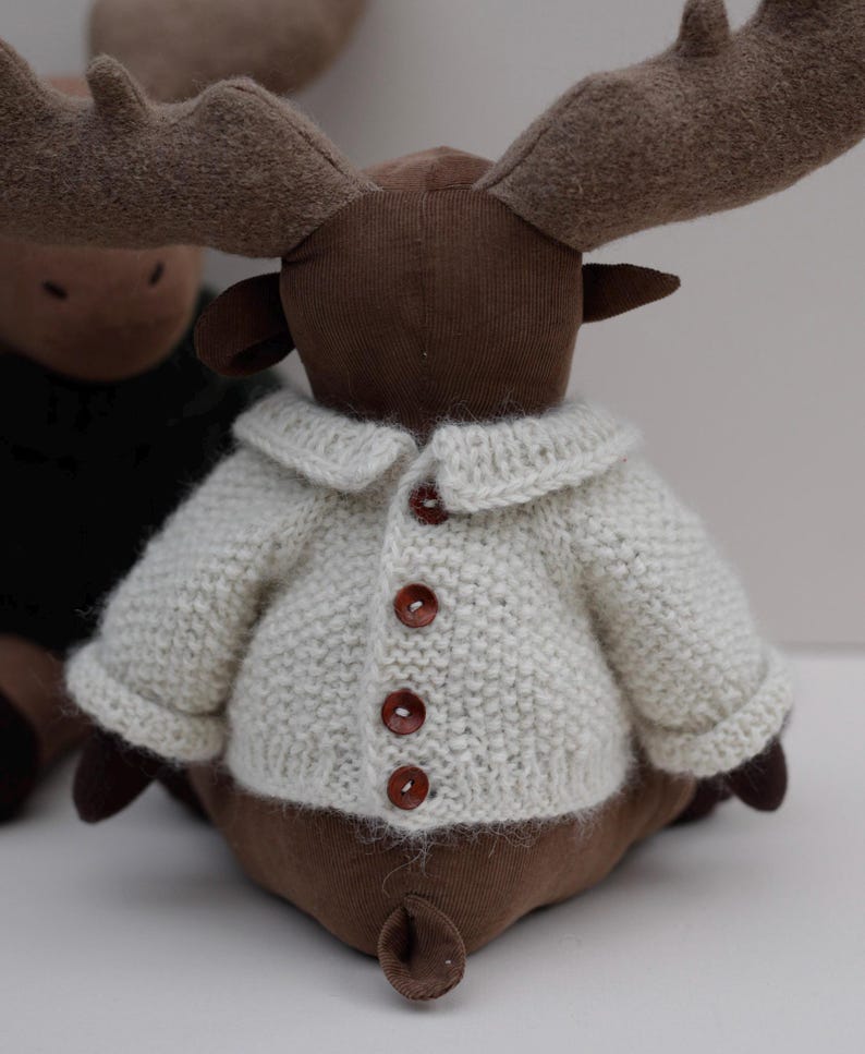 May include: A brown plush moose wearing a white knitted cardigan with brown buttons. The moose has large antlers and a brown face.