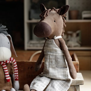 Puede incluir: Un caballo de peluche marr&oacute;n y beige con un mono de cuadros grises y blancos. El caballo est&aacute; sentado en un taburete de madera.