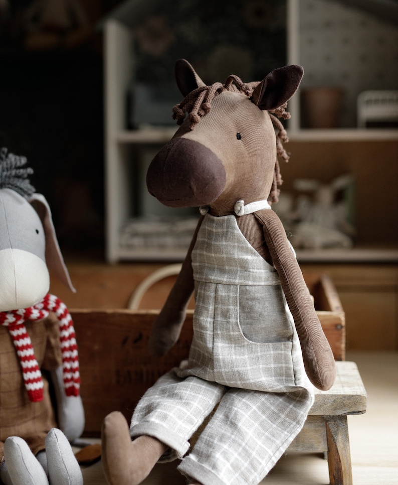 Puede incluir: Un caballo de peluche marr&oacute;n y beige con un mono de cuadros grises y blancos. El caballo est&aacute; sentado en un taburete de madera.