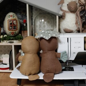 May include: Two brown plush platypus toys sitting side by side on a white surface. The platypus on the right is wearing a green and white spotted umbrella. The toys are in front of a white sewing machine with the number "b79" on the side.