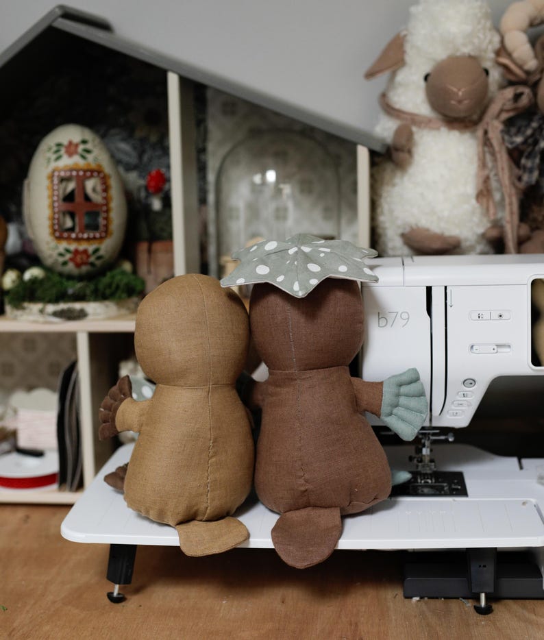 May include: Two brown plush platypus toys sitting side by side on a white surface. The platypus on the right is wearing a green and white spotted umbrella. The toys are in front of a white sewing machine with the number "b79" on the side.