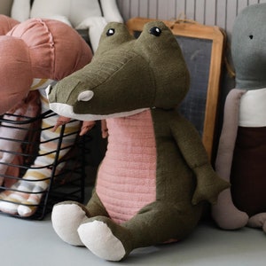 May include: A plush green and pink crocodile toy with white feet. The toy is sitting on a light gray surface.