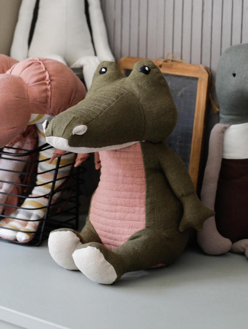 May include: A plush green and pink crocodile toy with white feet. The toy is sitting on a light gray surface.
