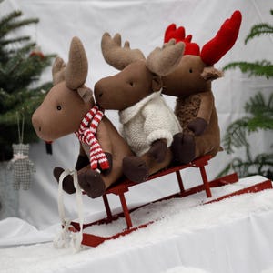 May include: Three brown plush moose with red antlers are riding on a red wooden sled. The moose in the front is wearing a white and brown striped scarf. The moose in the middle is wearing a white knitted sweater. The moose in the back is wearing a red and white striped scarf.