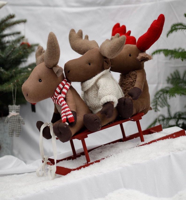 May include: Three brown plush moose with red antlers are riding on a red wooden sled. The moose in the front is wearing a white and brown striped scarf. The moose in the middle is wearing a white knitted sweater. The moose in the back is wearing a red and white striped scarf.