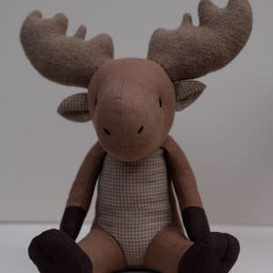 May include: A brown plush moose toy with large ears and a plaid pattern on its chest. The moose is sitting with its legs crossed and has a friendly expression.