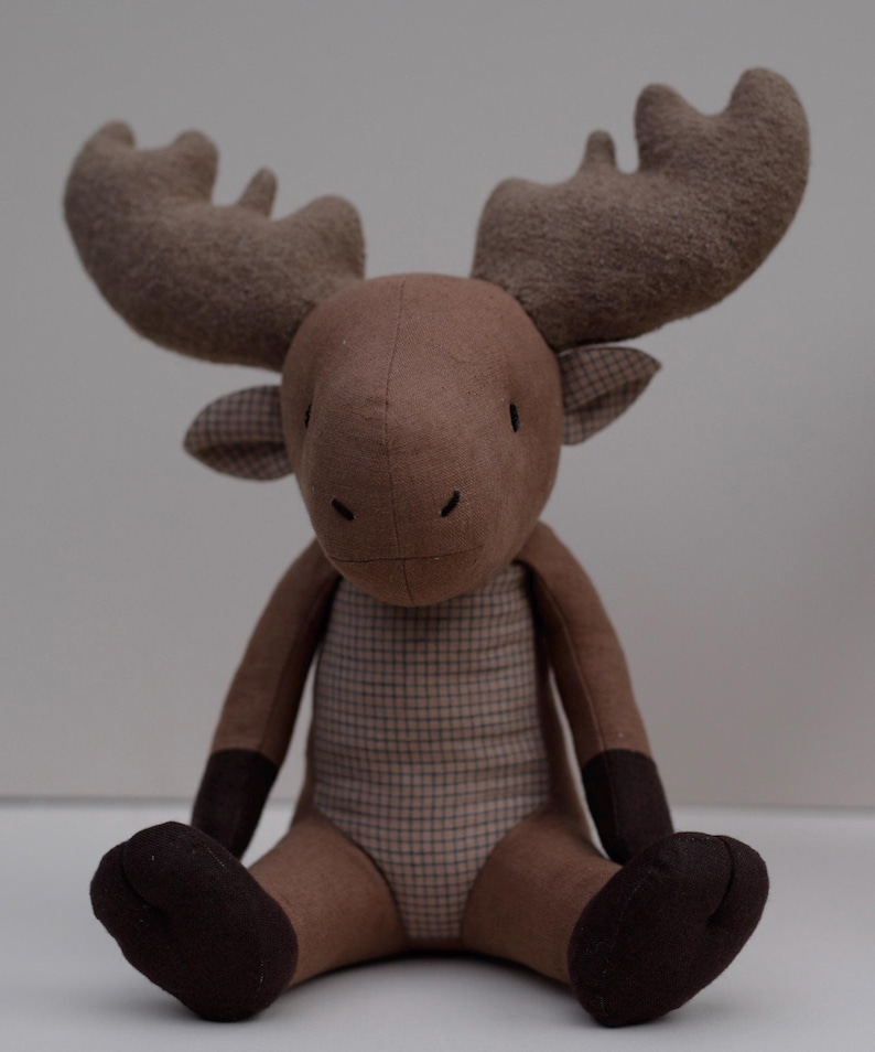 May include: A brown plush moose toy with large ears and a plaid pattern on its chest. The moose is sitting with its legs crossed and has a friendly expression.
