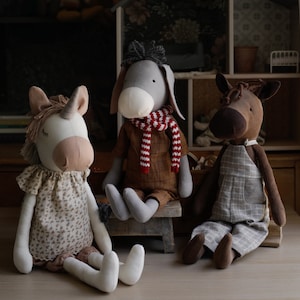 Puede incluir: Tres juguetes de peluche: un burro gris con una bufanda a rayas rojas y blancas, un caballo marr&oacute;n con un mono de cuadros y un unicornio blanco con un vestido de flores.