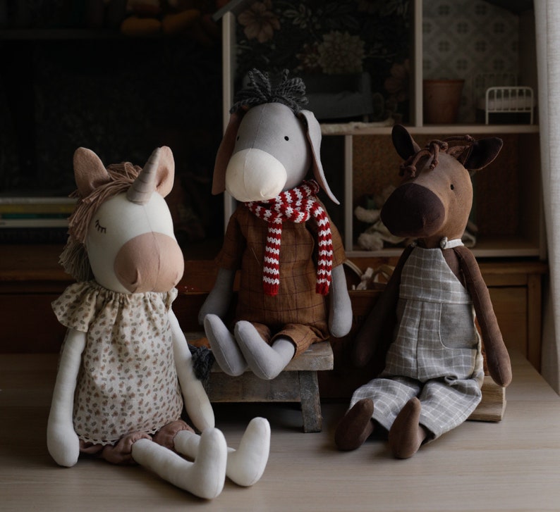 Puede incluir: Tres juguetes de peluche: un burro gris con una bufanda a rayas rojas y blancas, un caballo marr&oacute;n con un mono de cuadros y un unicornio blanco con un vestido de flores.