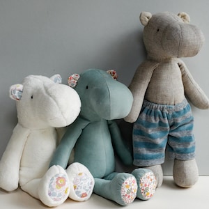 May include: Three plush hippopotamus toys, one white, one teal blue, and one gray. The white and teal blue hippos have floral fabric on their feet. The gray hippo is wearing blue and gray striped pants.