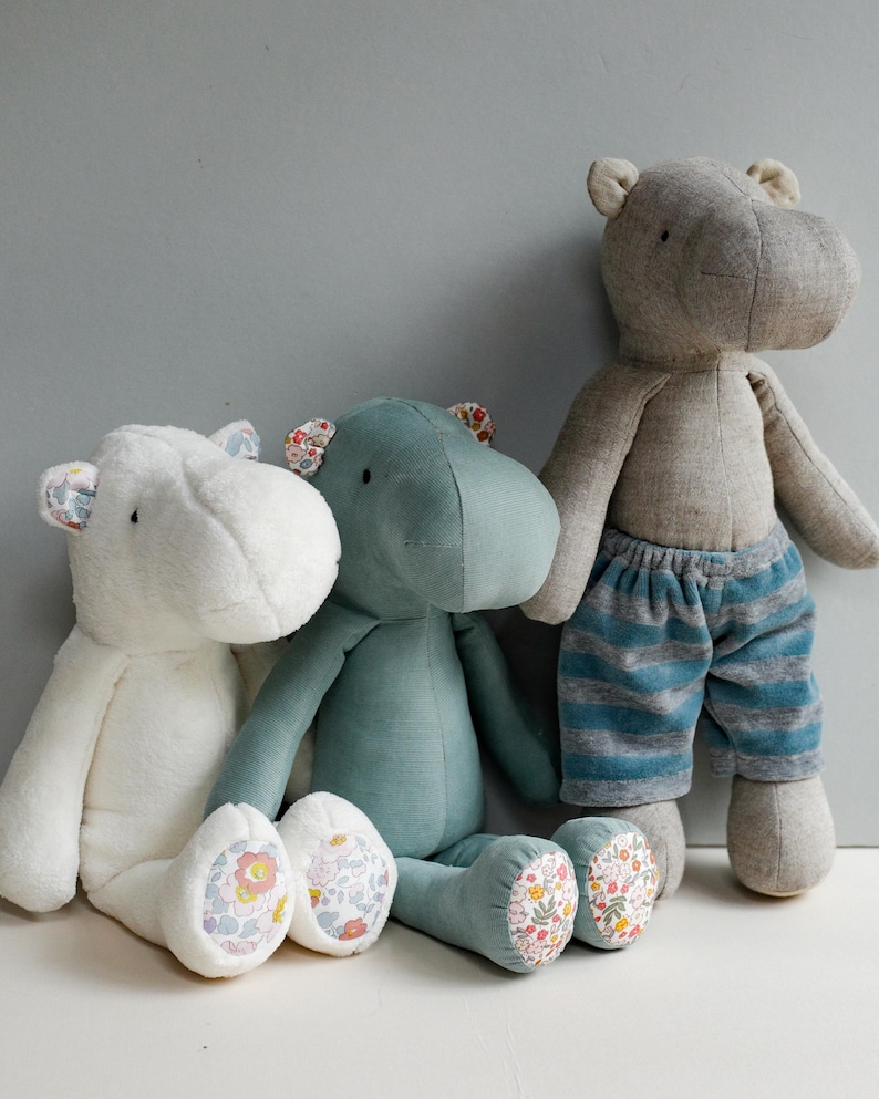 May include: Three plush hippopotamus toys, one white, one teal blue, and one gray. The white and teal blue hippos have floral fabric on their feet. The gray hippo is wearing blue and gray striped pants.