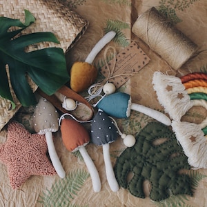 Puede incluir: Un conjunto de juguetes de setas de fieltro hechos a mano con un arco&iacute;ris, una hoja y una estrella. Las setas son de varios colores, incluyendo marr&oacute;n, gris, azul verdoso y naranja. El arco&iacute;ris est&aacute; hecho de hilo y tiene flecos. La hoja es verde y la estrella es rosa.