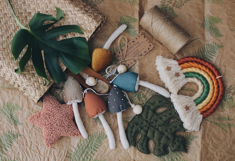 Puede incluir: Un conjunto de juguetes de setas de fieltro hechos a mano con un arco&iacute;ris, una hoja y una estrella. Las setas son de varios colores, incluyendo marr&oacute;n, gris, azul verdoso y naranja. El arco&iacute;ris est&aacute; hecho de hilo y tiene flecos. La hoja es verde y la estrella es rosa.