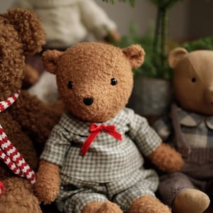 May include: Three plush teddy bears, one wearing a plaid shirt and red bow tie, another with a white sweater, and a third with a red and white striped scarf. The bears are sitting on a wooden surface in front of a small potted Christmas tree.