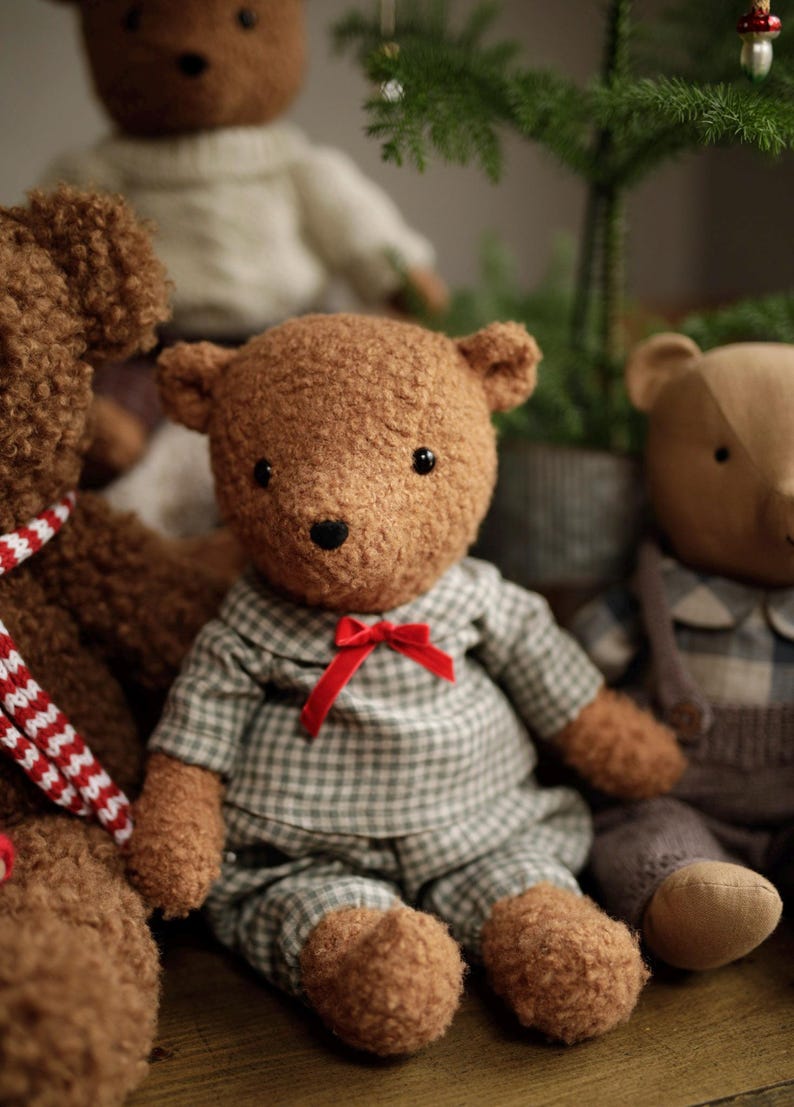 May include: Three plush teddy bears, one wearing a plaid shirt and red bow tie, another with a white sweater, and a third with a red and white striped scarf. The bears are sitting on a wooden surface in front of a small potted Christmas tree.