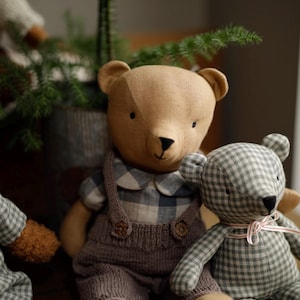 May include: Two plush teddy bears, one brown with a plaid shirt and brown overalls, and one green and white plaid bear with a pink ribbon. The bears are sitting on a wooden surface.