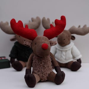 May include: Three plush reindeer toys, one with red antlers and a brown fuzzy body, and two with brown antlers and white sweaters. The reindeer are sitting on a white surface.