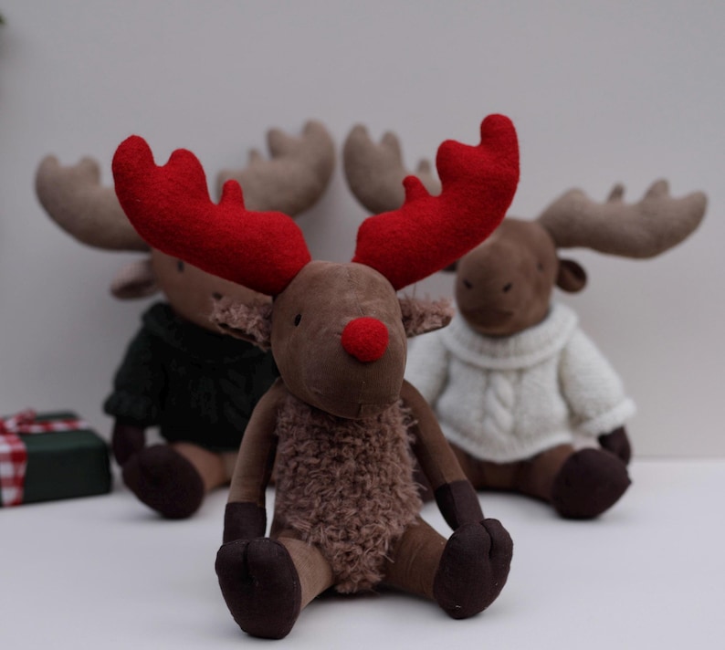 May include: Three plush reindeer toys, one with red antlers and a brown fuzzy body, and two with brown antlers and white sweaters. The reindeer are sitting on a white surface.
