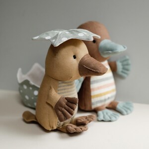 May include: Two plush toy platypuses, one wearing a green and white polka dot umbrella. The platypus on the left is brown and white striped, while the platypus on the right is brown and has a blue beak.
