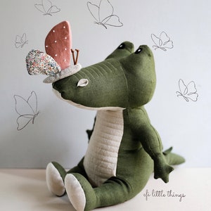 May include: A green plush crocodile toy with a pink and white butterfly on its head. The crocodile is sitting on a white surface with a light blue background. The butterfly has a floral pattern and is sitting on the crocodile's head. The crocodile has a stitched design on its back and a white belly. The image has the text "efi little things" in the bottom right corner.