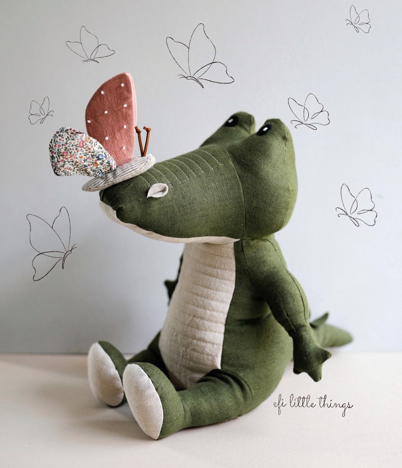 May include: A green plush crocodile toy with a pink and white butterfly on its head. The crocodile is sitting on a white surface with a light blue background. The butterfly has a floral pattern and is sitting on the crocodile's head. The crocodile has a stitched design on its back and a white belly. The image has the text "efi little things" in the bottom right corner.