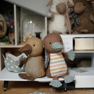 May include: Two brown and beige plush toy platypuses with embroidered eyes and beaks. The platypus on the left has a striped pattern on its body. The platypus on the right has a striped pattern on its chest and a light blue beak. The platypuses are sitting on a white surface.