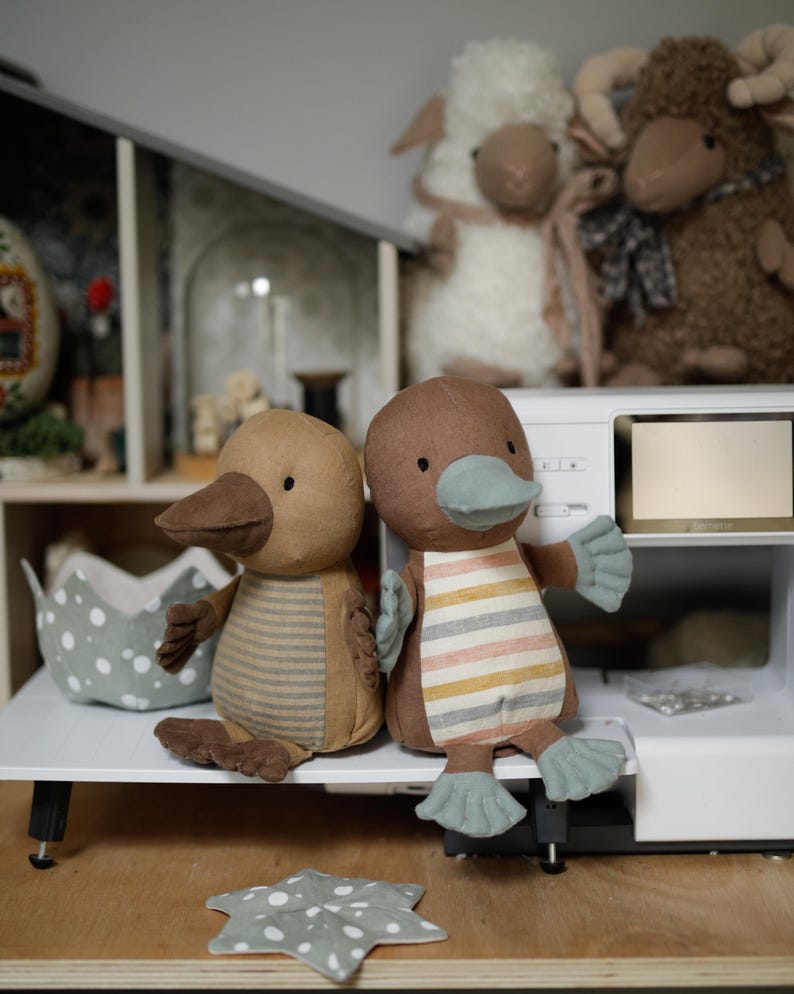 May include: Two brown and beige plush toy platypuses with embroidered eyes and beaks. The platypus on the left has a striped pattern on its body. The platypus on the right has a striped pattern on its chest and a light blue beak. The platypuses are sitting on a white surface.