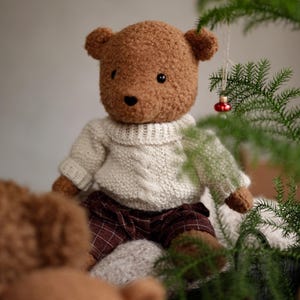 May include: A brown teddy bear wearing a white knitted sweater and plaid pants sits near a green plant with a red ornament.